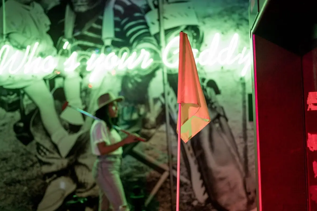 A person in a hat stands in front of a neon sign reading "Who's your Caddy" at one of the best mini golf places in the Bay Area. The golf-themed indoor area features vintage black-and-white golfing images, and a small flag is seen in the foreground. The lighting is predominantly green and red.
