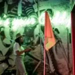 A person in a hat stands in front of a neon sign reading "Who's your Caddy" at one of the best mini golf places in the Bay Area. The golf-themed indoor area features vintage black-and-white golfing images, and a small flag is seen in the foreground. The lighting is predominantly green and red.