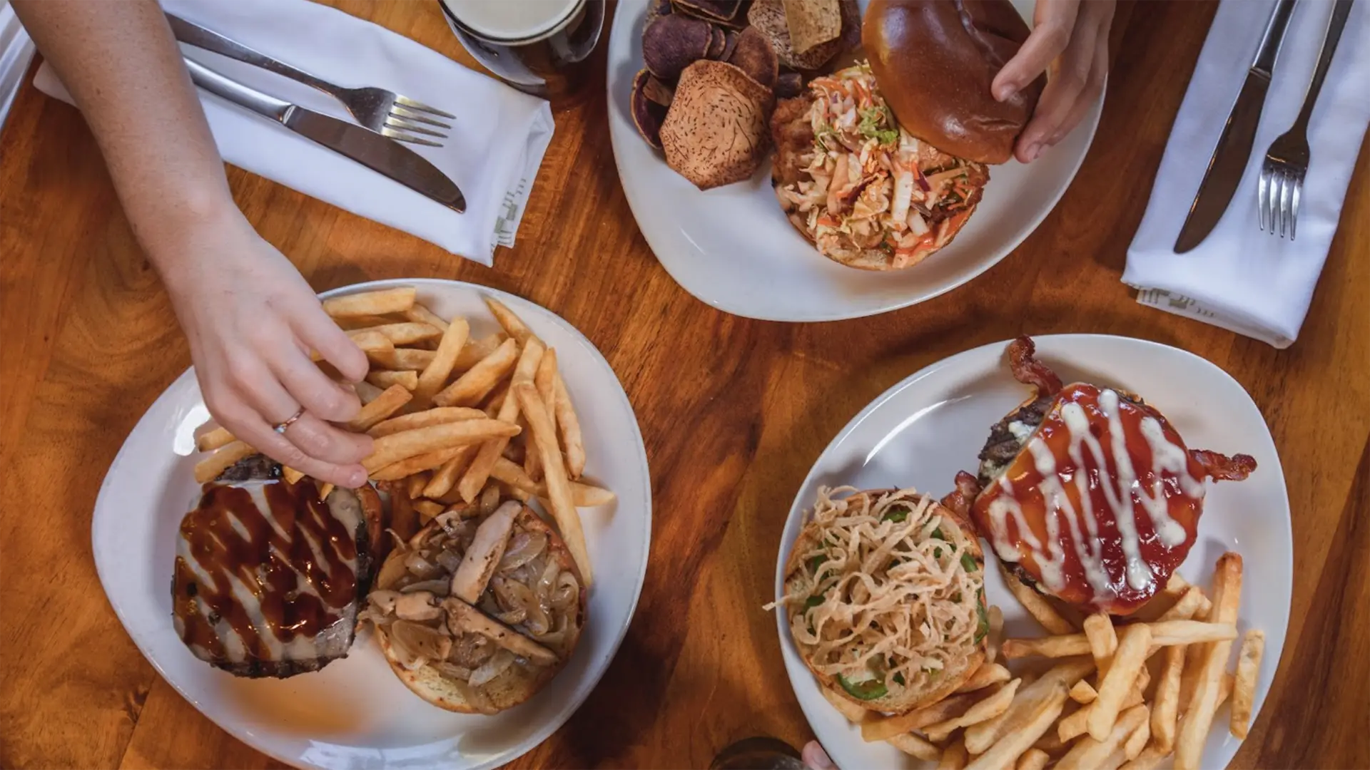 Overhead view of a wooden table with three plates of food, perfect for the best lunch on Kauai's North Shore. Each plate boasts a burger and fries, topped uniquely with coleslaw, crispy onions, or barbecue sauce. Hands eagerly reach for the delectable spread.