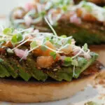 A close-up of two pieces of toasted bread, each topped with slices of avocado, diced tomatoes, microgreens, and a sprinkle of seasoning. The bread is set on a white plate with scattered seasoning visible around the edges, reminiscent of the best Hawaiian food found in Kauai.