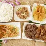 An assortment of dishes including white rice, shredded meat, fried shrimp, breaded cutlets, grilled meats, and vegetable salad served on rectangular white plates, arranged on a sandy surface with a pineapple and purple flowers as decoration. This spread showcases the best Hawaiian food Kauai has to offer.
