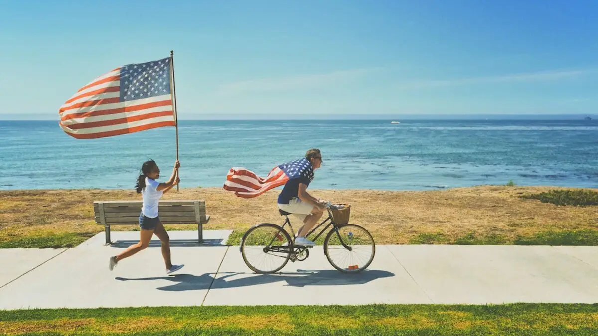 A person rides a bicycle along a seaside path, with an American flag waving from the back. Another person runs alongside, energetically holding a large American flag. The ocean and blue sky provide a scenic backdrop—one of the best things to do in the Bay Area Labor Day Weekend.