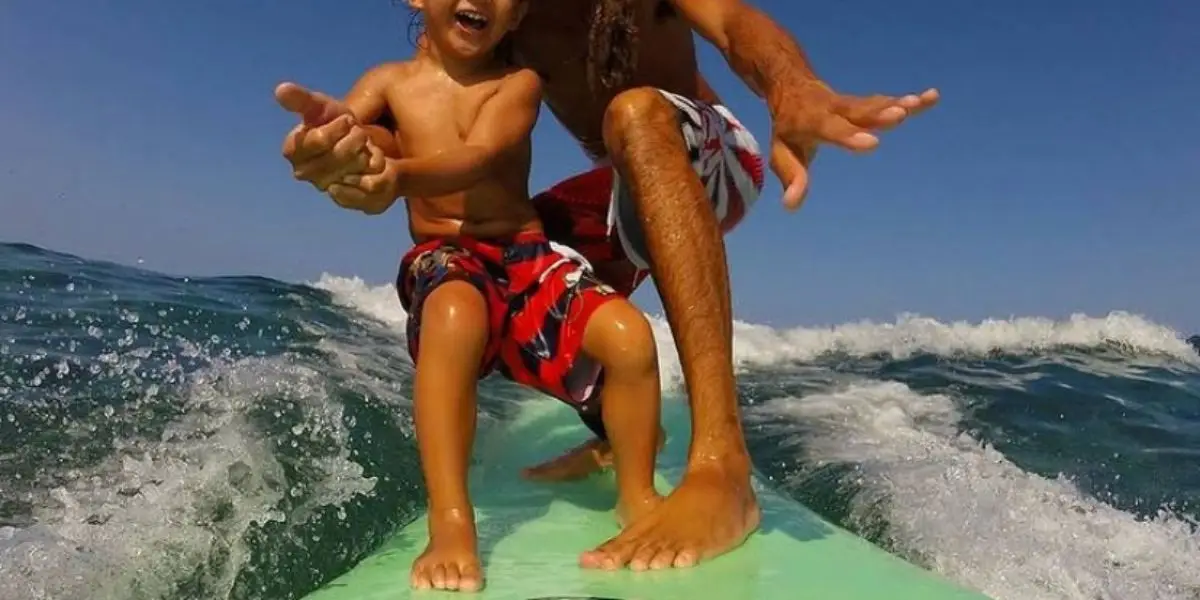 A child and an adult are surfing together on a green surfboard, riding a wave in the ocean. Both are wearing red and black patterned shorts and appear to be enjoying the moment under a clear, blue sky. The adult, who might just be from the top surf lessons on the Big Island, has their arm around the child, guiding them.