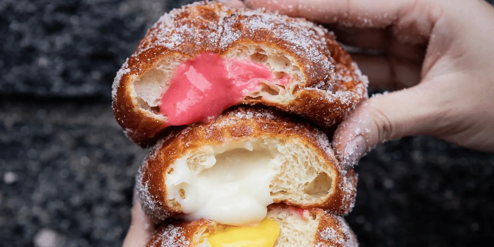 A close-up shot of a person holding three halves of sugar-coated, filled pastries stacked on top of each other. The pastries are filled with pink, white, and yellow cream, respectively, and have a flaky, doughy texture – truly some of the best desserts Maui Wailea has to offer.