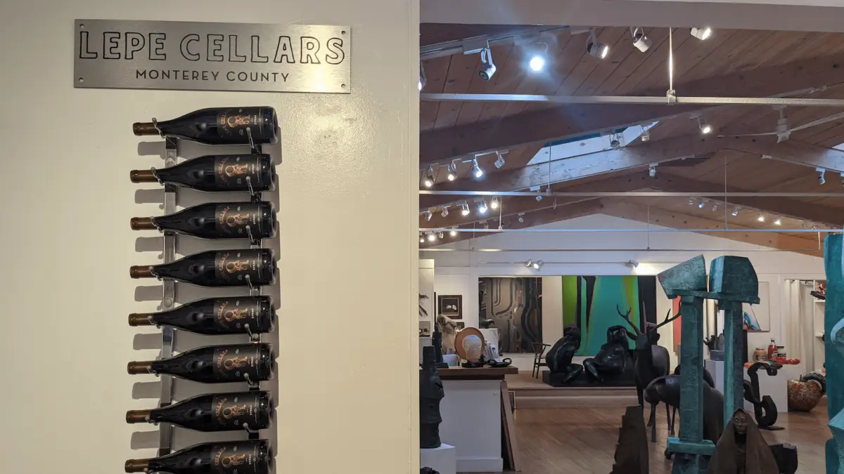 A display of nine wine bottles mounted on the wall under a "Lepe Cellars Monterey County" sign is seen on the left. On the right, an art gallery with various sculptures and artworks scattered throughout the space evokes the charm found in Carmel Wine Tasting Rooms.