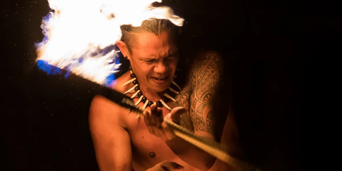 A shirtless man performs a fire dance against a dark background. He grips a flaming staff and wears a tribal necklace, with tribal tattoos on his arm. The flames are bright, illuminating his determined expression, reminiscent of ancient Hawaiian luau history.