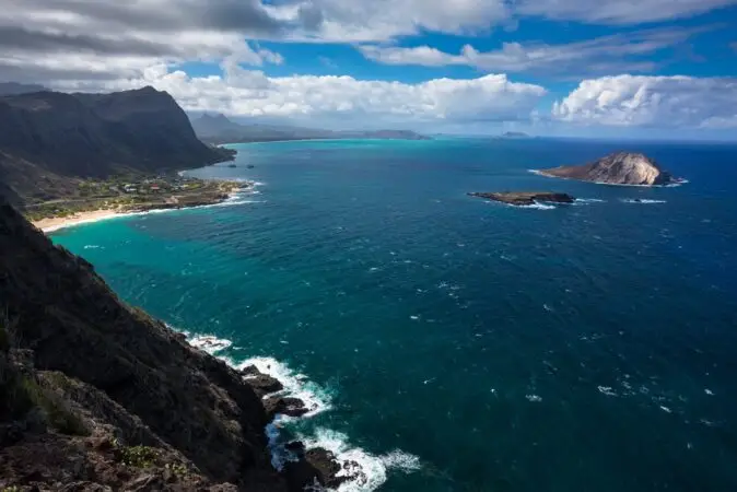A breathtaking coastal view showcasing the vibrant turquoise sea of Makapuu Beach Oahu merging with the deeper blue ocean. A stretch of rocky coastline and small islands is visible under a partly cloudy sky, with a distant mountain range adding depth to this stunning landscape.