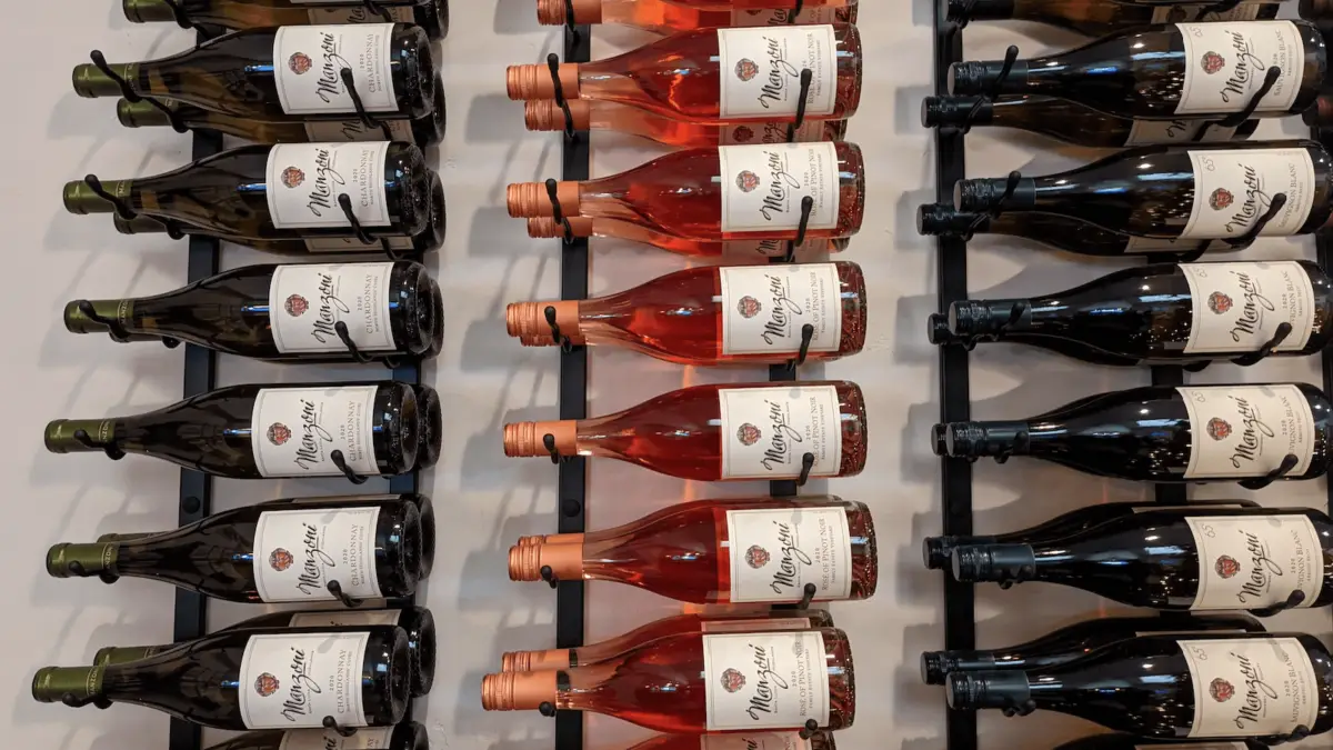 A wall-mounted wine rack holding multiple bottles of wine is displayed. The rack has three columns with various bottles arranged horizontally. The wine colors range from red and rosé to white, each with a visible label, reminiscent of selections found in Carmel Wine Tasting Rooms.