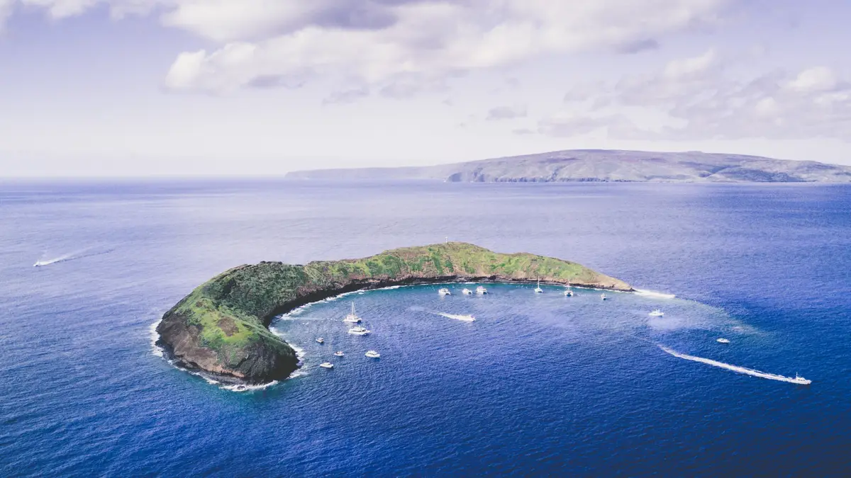 Aerial view of a small, crescent-shaped island, one of the top attractions in Maui, surrounded by clear blue ocean waters. Numerous boats are anchored around the island, which is covered in lush green vegetation. The sky is partly cloudy, and distant landmasses are visible on the horizon.