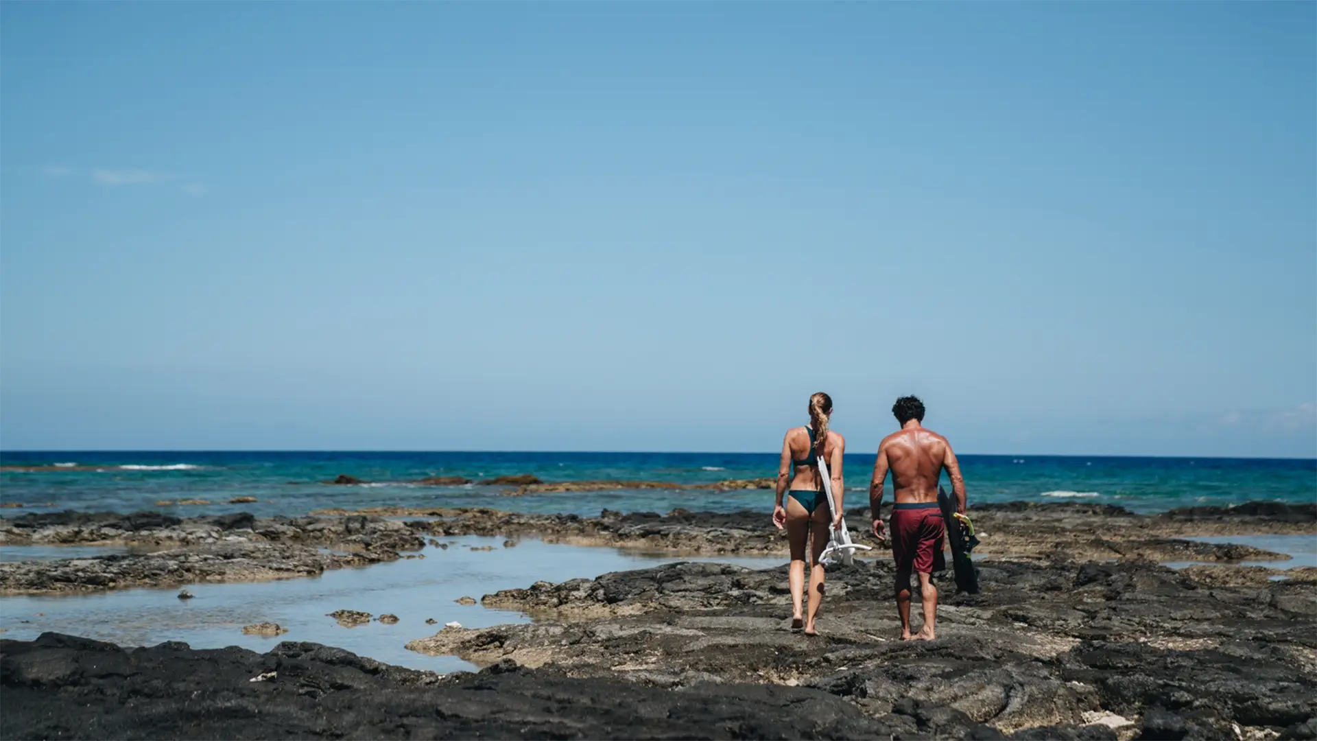 Two people in swimwear walk along a rocky shoreline towards the ocean. The clear blue sky and calm sea create a serene beach scene. Both individuals carry snorkeling gear, suggesting they're heading for a swim or dive, perhaps after staying at one of the best romantic hotels on the Big Island.