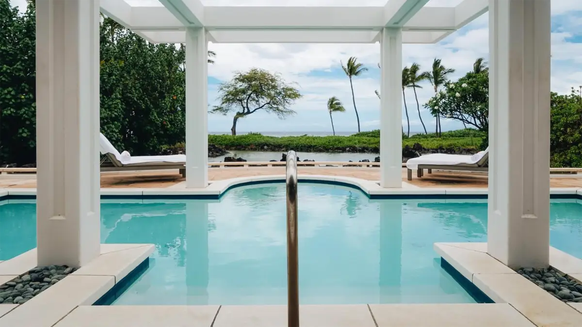 A luxurious outdoor pool with clear blue water is framed by white columns and tropical trees. Lounge chairs with white towels are positioned at both sides of the pool. Beyond the pool, lush greenery and palm trees overlook the calm ocean under a partly cloudy sky, typical of the best Big Island luxury hotels.