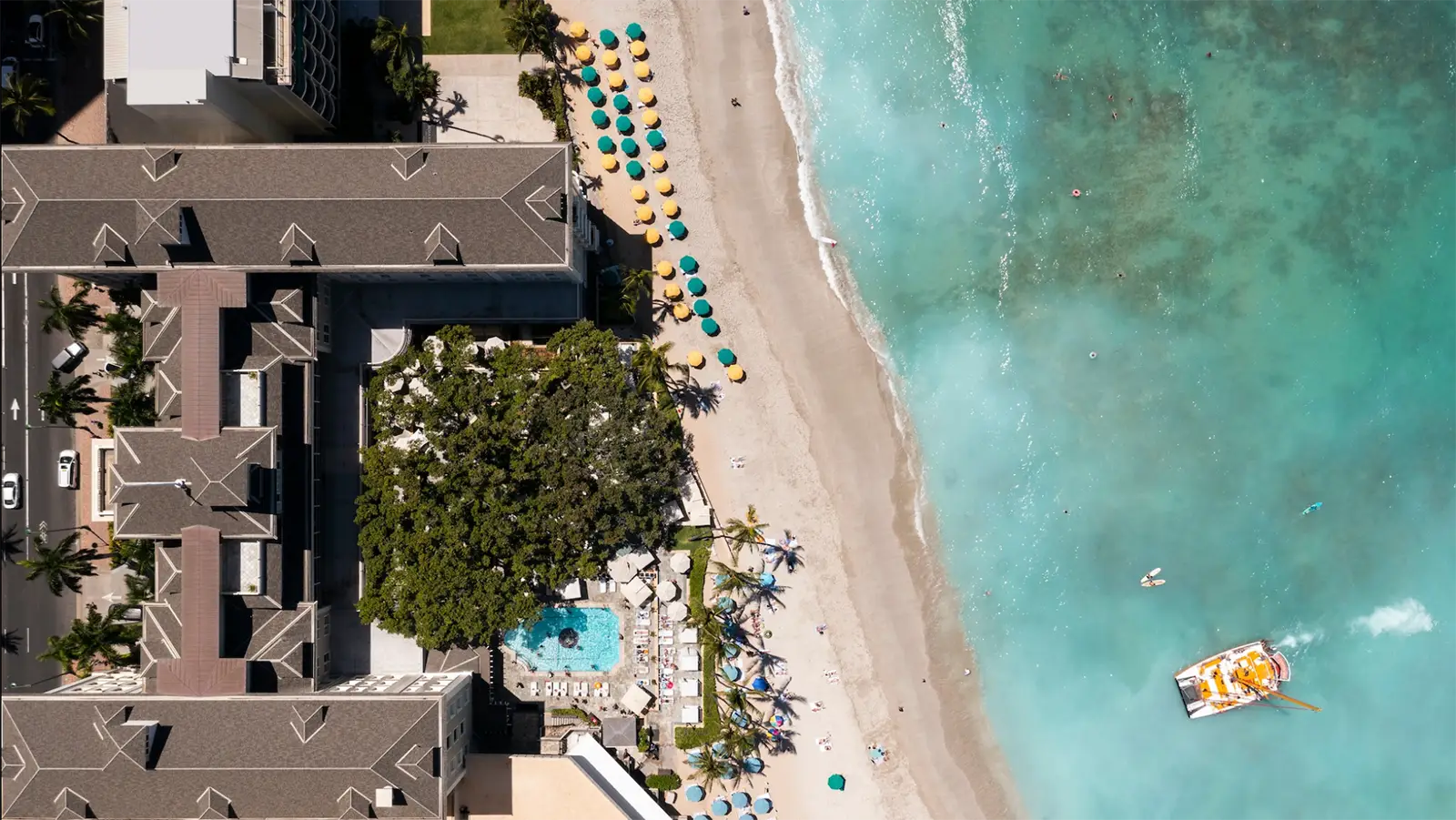 Aerial view of a beachfront resort, one of the best oceanfront hotels Oahu offers, with a turquoise pool surrounded by lounge chairs and umbrellas. The sandy beach is lined with yellow and green umbrellas, leading to clear blue ocean water where a boat with a yellow canopy is anchored.