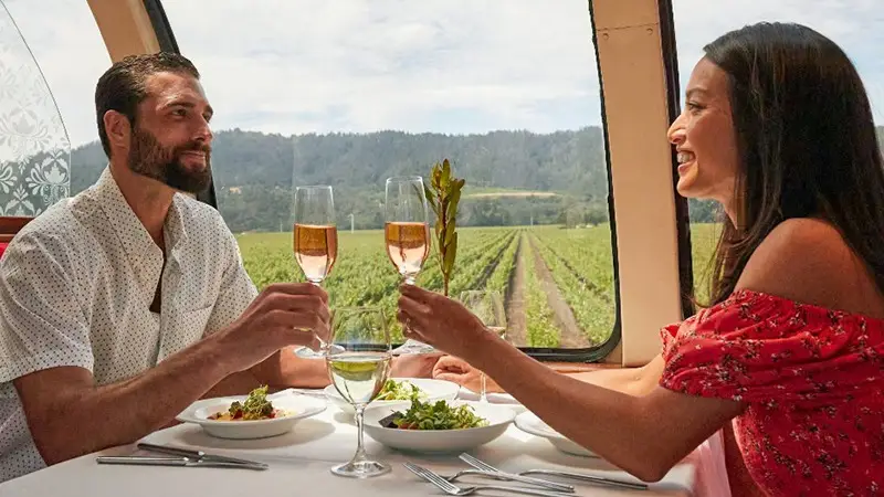A man and woman toast with glasses of wine while dining on a luxurious train with large windows offering a scenic view of a vineyard. The man wears a white shirt, and the woman wears a red dress. They are smiling and enjoying one of the best things to do in Wine Country in January.