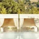 A luxurious outdoor pool area with mist rising from the water. In the background are cabanas draped with light fabric, providing shade and seating with cushions. Beyond the cabanas, green trees and a mountainous landscape complete the serene setting at one of Napa's Best Spa Wellness Hotels.
