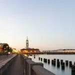 A waterfront view of the Ferry Building in San Francisco at dusk. The scene features city lights twinkling along the pier, calm waters reflecting city structures, and a clear sky. Buildings, including the Harbor Court Hotel, line the promenade with the iconic clock tower standing prominently.
