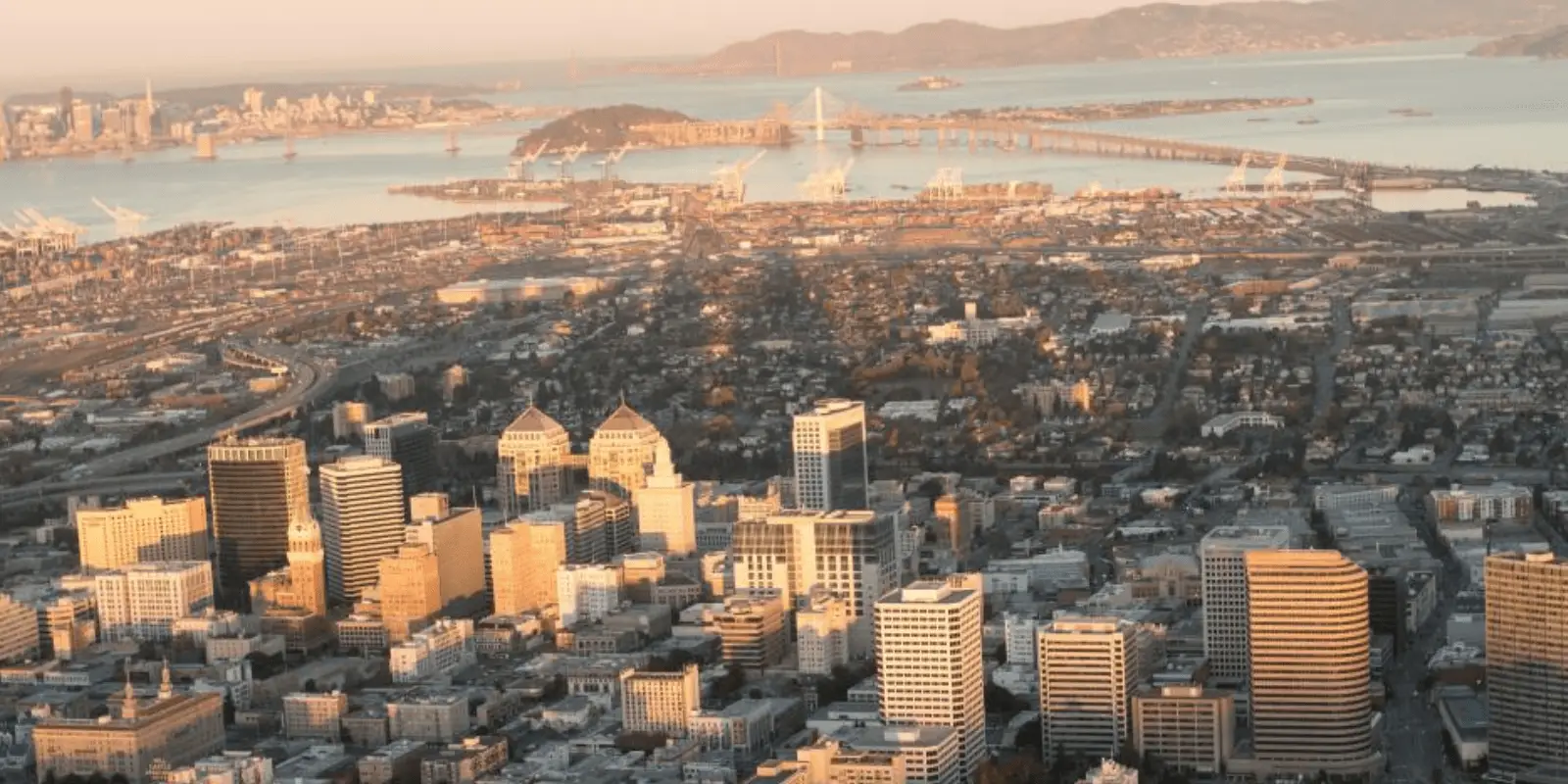 Aerial view of a cityscape during sunset. Tall buildings and skyscrapers are seen in the foreground, with roads and bridges extending towards a body of water in the background. Hills and distant landscapes are visible under a warm, golden sky—a perfect moment to reflect on the Best Things to Do in the East Bay November 2021.