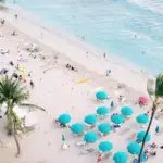 Aerial view of a beach filled with people, umbrellas, and lounge chairs. The sandy shore is dotted with teal umbrellas and beachgoers enjoying the sun. The turquoise ocean waves gently lap against the coastline, with a few swimmers visible in the water. Palm trees add a tropical touch, completing an ideal scene for those staying at best value hotels in Oahu.