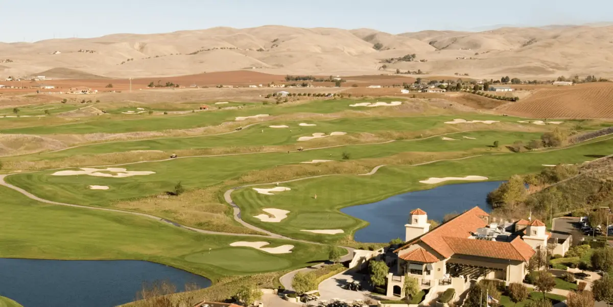 A sprawling golf course set against rolling beige hills, featuring well-manicured green fairways, multiple sand bunkers, and water hazards. In the foreground is a large clubhouse with a terracotta roof, surrounded by trees and pathways. It's one of the best golfing destinations in the East Bay.