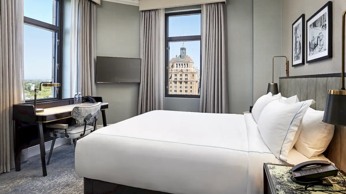 A modern hotel room with gray walls featuring a large bed with white linens, a wall-mounted TV, a work desk with a chair, and two windows providing a view of a historic building outside. Two framed pictures are hung on the wall above the bed, perfect for guests exploring "The Ultimate Guide to Sacramento.
