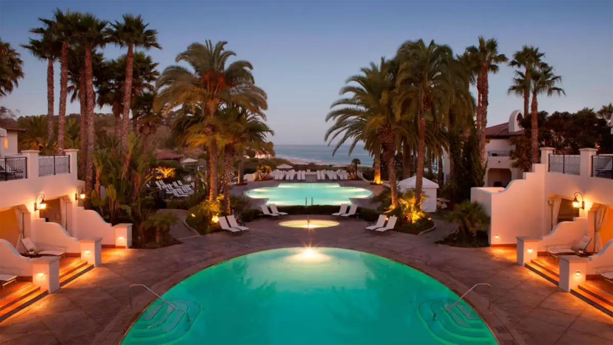 A luxurious resort pool area at twilight, featuring two elegant pools surrounded by palm trees, lounge chairs, and umbrellas. The pools are illuminated, creating a serene atmosphere that makes this South Bay hotel the best romantic getaway in Santa Barbara. In the background, a small path leads towards the ocean horizon.