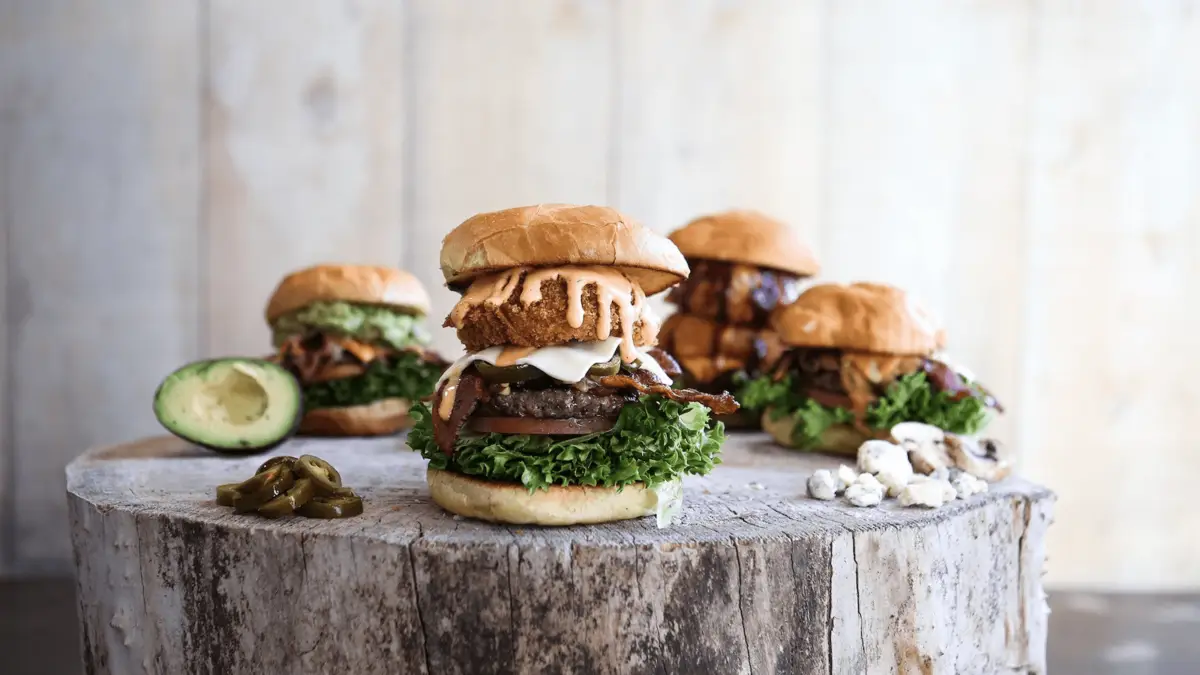 A close-up of a gourmet burger on a wooden surface, topped with lettuce, cheese, crispy onion rings, and sauce. Surrounding the main burger are three other varied burgers, sliced avocado, jalapeño slices, and crumbled blue cheese—all making this the best lunch on Oahu against a rustic background.