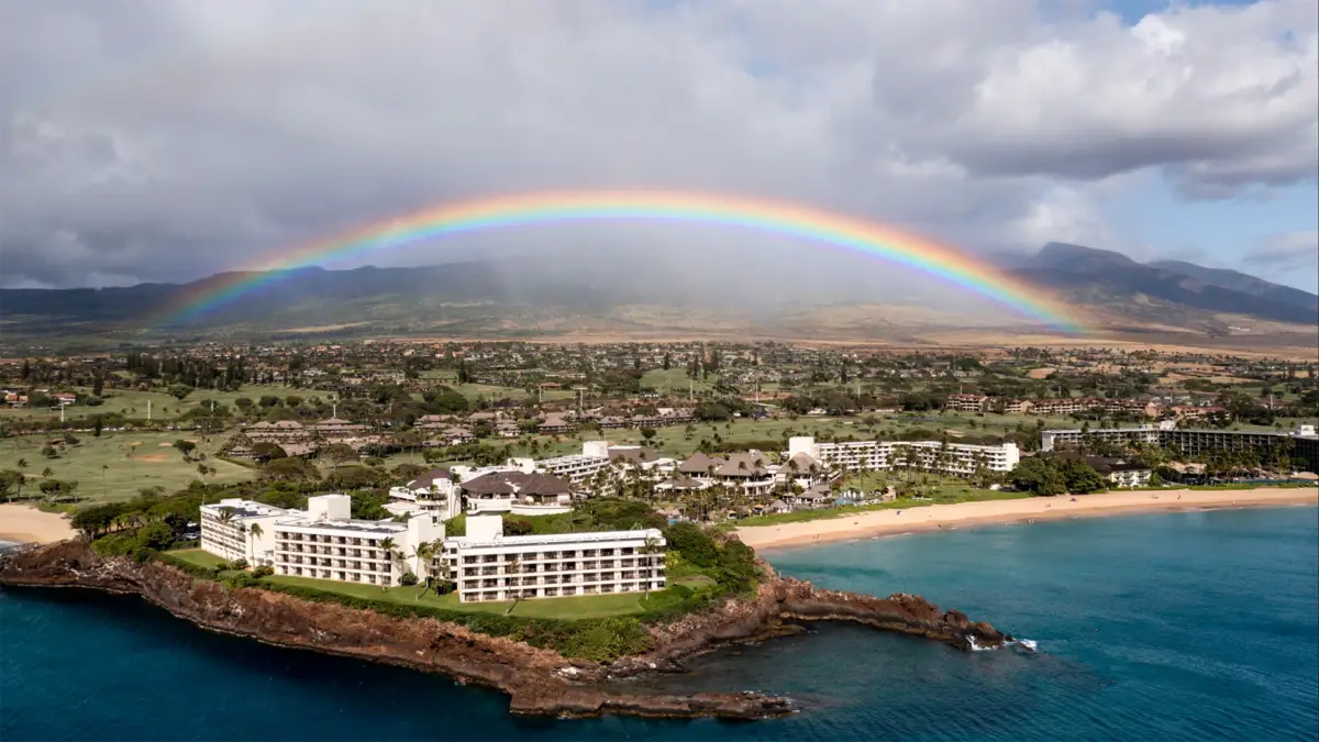 Aerial view of a coastal resort with white buildings on a peninsula, surrounded by blue ocean water and sandy beaches. A vibrant rainbow arches across the sky over the lush green landscape and mountains in the background under a cloudy sky—truly one of the best group hotels Maui has to offer.