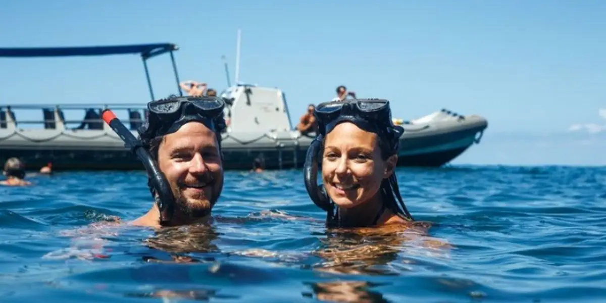 Two snorkelers, a man and a woman, smile at the camera while bobbing in the ocean. They wear black snorkel masks. A boat with other people is anchored nearby under a clear blue sky, enjoying one of the best snorkeling tours Kauai has to offer.