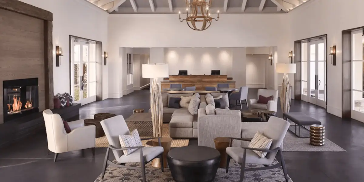 A modern hotel lobby featuring contemporary decor, ideal for the best group getaway hotels in Sonoma. The area has plush armchairs, a cozy sofa, wooden coffee tables, and light fixtures. Behind the seating area, a large wooden reception desk with three computers is visible. The room boasts high ceilings and large windows.