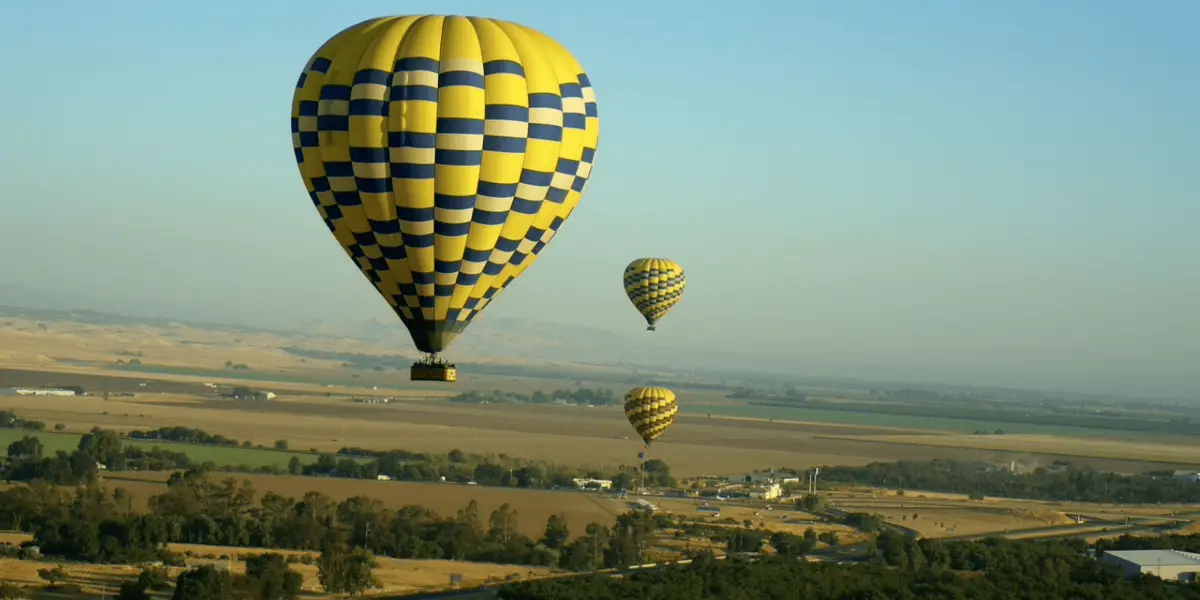 A scenic view in Sonoma Wine Country shows three hot air balloons in flight over a vast landscape of fields and farmlands. The balloons are yellow and blue checkered, floating serenely above the green and golden terrain under a clear blue sky, capturing the essence of summer adventures.