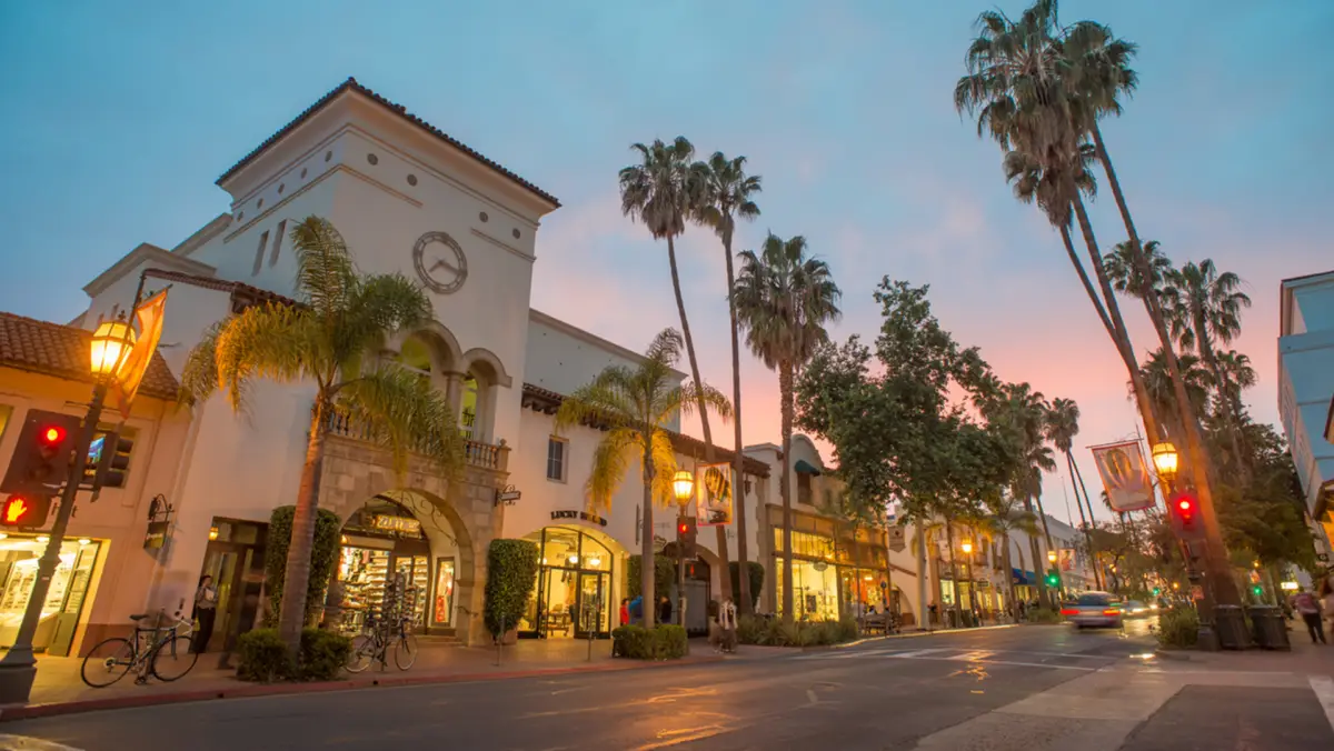 A charming street scene at sunset featuring a historic building with a clock tower surrounded by tall palm trees. The street, one of the best places to visit in Santa Barbara, is lined with shops and illuminated by street lamps, with a few people and bicycles visible under the evening sky.
