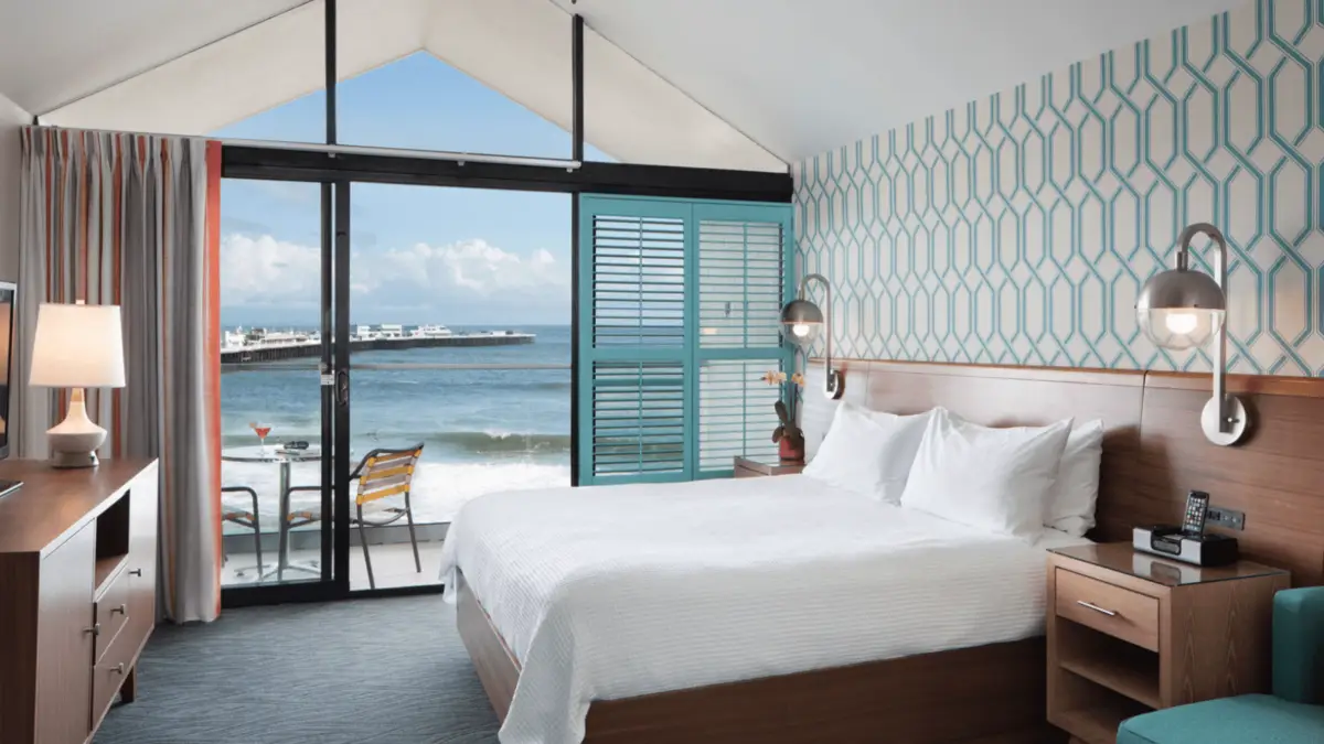 A modern hotel room with a king-sized bed, wooden furniture, and a geometric patterned accent wall. The room leads out to a balcony with a table and chairs, overlooking a scenic ocean view with waves and a pier in the distance—one of the Four Santa Cruz Hotels to put on your Radar.
