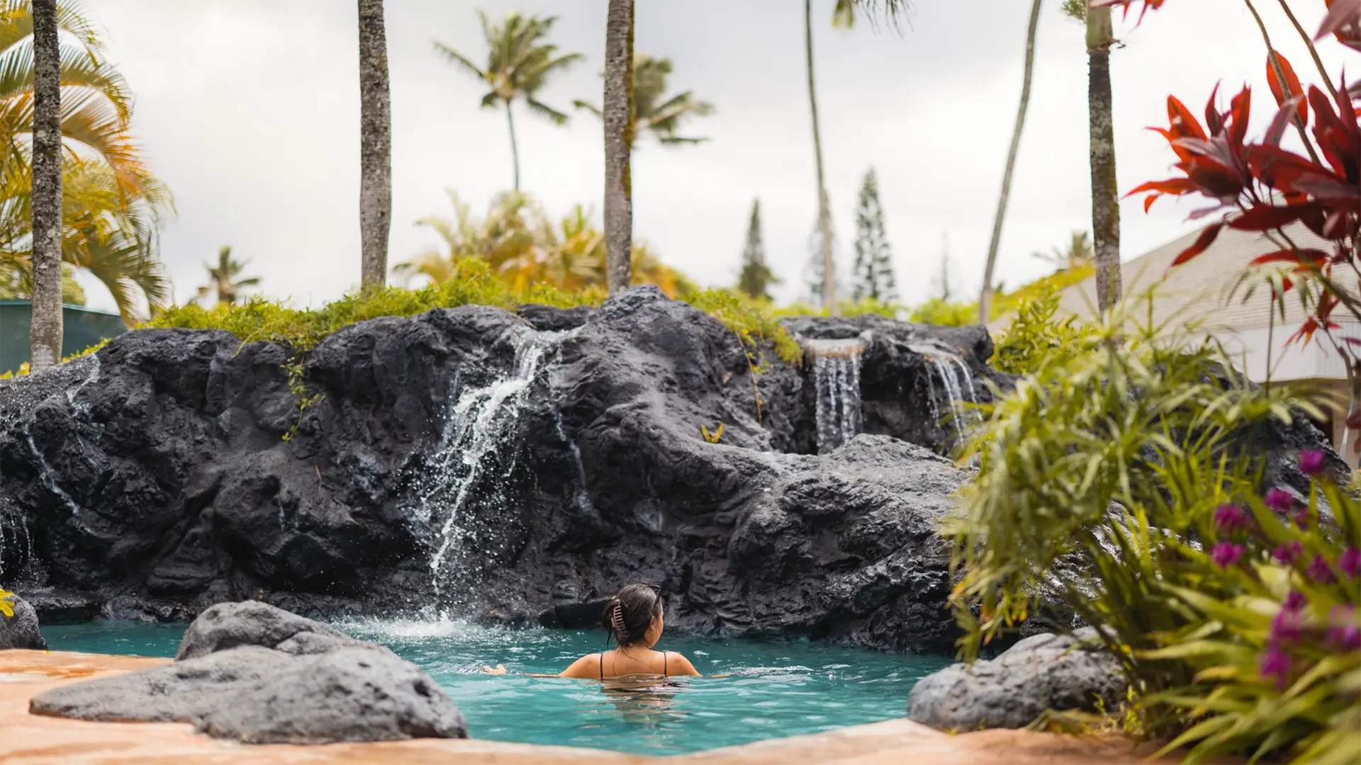 A person is seen from behind swimming in a serene pool surrounded by large rocks with small waterfalls cascading down. Palm trees and lush tropical plants create a picturesque background under a cloudy sky, reminiscent of the tranquil settings at the best family hotels in Kauai.