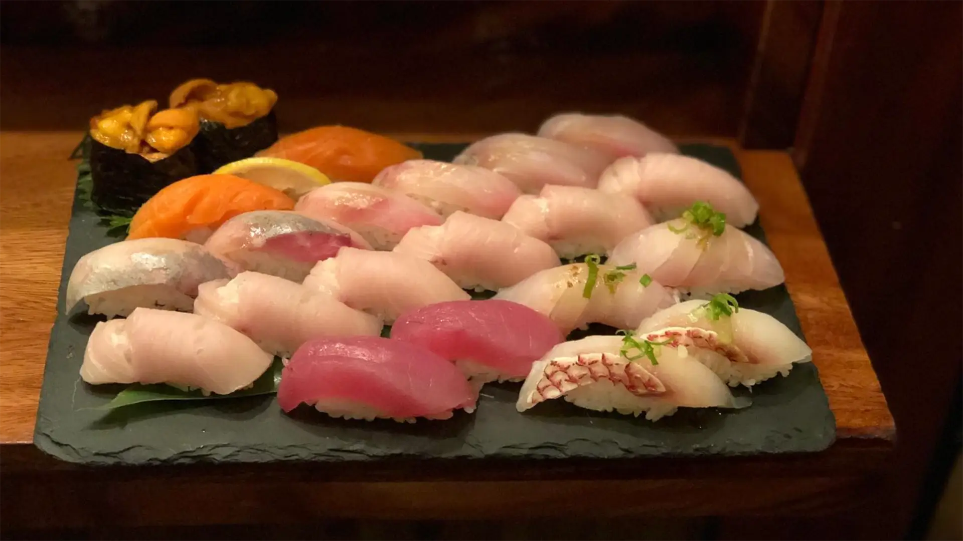 A display of assorted sushi pieces on a black slate platter. The best sushi in Kauai includes various types like tuna, salmon, and white fish, each beautifully crafted and garnished with green onions, radish, or roe. The platter also contains a piece of uni (sea urchin).