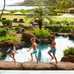 Three children in swimsuits, one girl and two boys, walk in a line along a stone edge of a swimming pool surrounded by lush greenery at one of the best family hotels Kauai offers. The background features a grassy area with lounge chairs and umbrellas, with a view of water and hills in the distance.