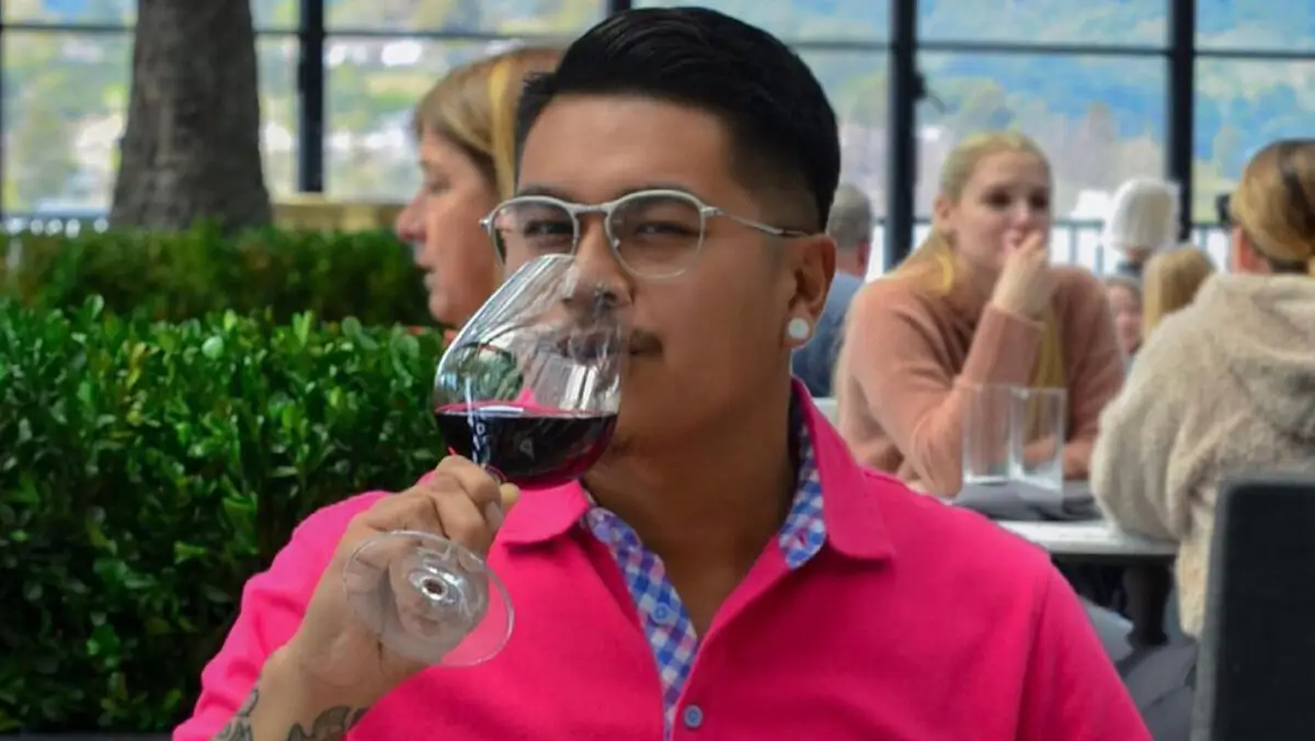 A person wearing glasses, a pink shirt, and sporting a tattoo on their forearm, is sitting at an outdoor restaurant in the Bay Area, savoring a glass of red wine. The background shows other diners, greenery, and a scenic view—perfect for Instagram.
