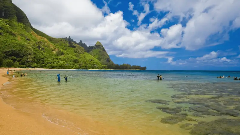A scenic beach in Kaua'i with clear blue water and a sandy shore is framed by green, rocky cliffs under a partly cloudy sky. People are wading in the water and swimming near the shore, enjoying the natural beauty of this hidden gem.