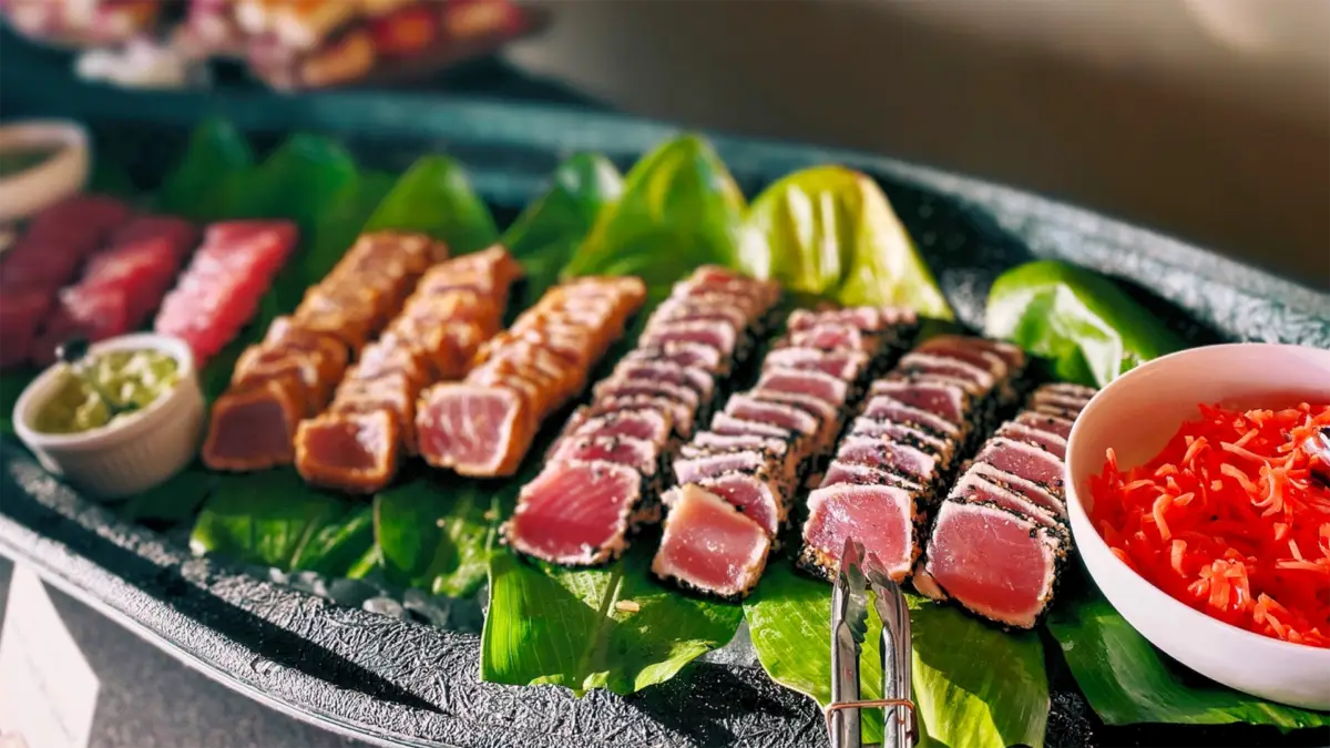 A platter of assorted sashimi is artfully arranged on fresh green leaves. The varieties include seared tuna, sesame-crusted tuna, and traditional raw tuna slices. A small bowl of bright red pickled ginger and a container of wasabi sit nearby—offering the best lunch on the Big Island.