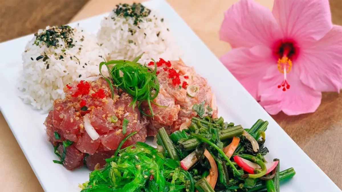 A rectangular white plate holds a variety of foods, including two scoops of white rice, two types of the best poke on the Big Island (diced raw fish with garnishes and sauce), and a serving of seaweed salad with vegetables. A pink hibiscus flower sits to the right side of the plate.
