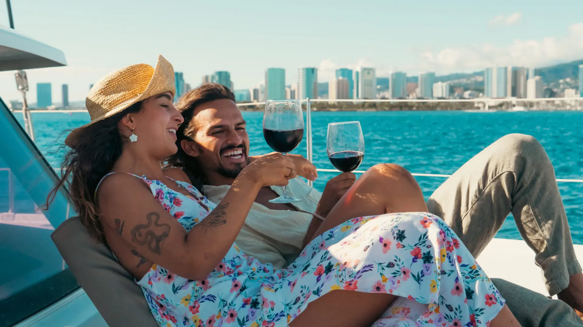 A couple relaxes on a boat, smiling and clinking wine glasses while enjoying the view of a city skyline across the water. The woman wears a floral dress and straw hat, and the man has a beard and wears a light shirt. It's a sunny day with clear blue skies—the best sunset cruise Oahu has to offer.