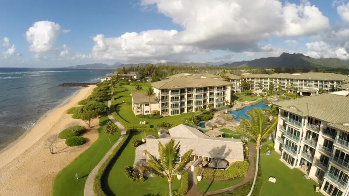 Aerial view of a coastal resort with beige buildings surrounded by lush green lawns and palm trees. A winding path leads to a sandy beach on the left. There's a pool area with blue waters near the center, and the backdrop features mountains under a partly cloudy sky, showcasing one of the best family hotels in Kauai.