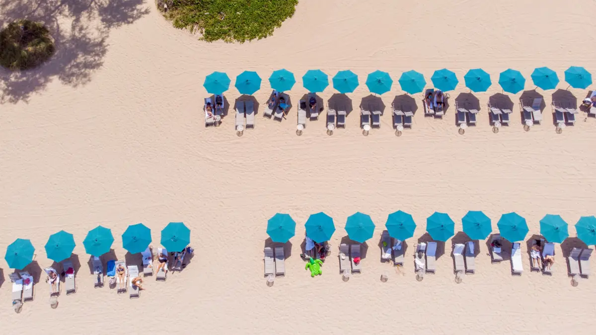 Aerial view of a sandy beach with two neat rows of blue umbrellas and sun loungers at the best group getaway hotel on Big Island. People are relaxing under the umbrellas, enjoying the sunny day. Green shrubbery is visible at the upper part of the image.