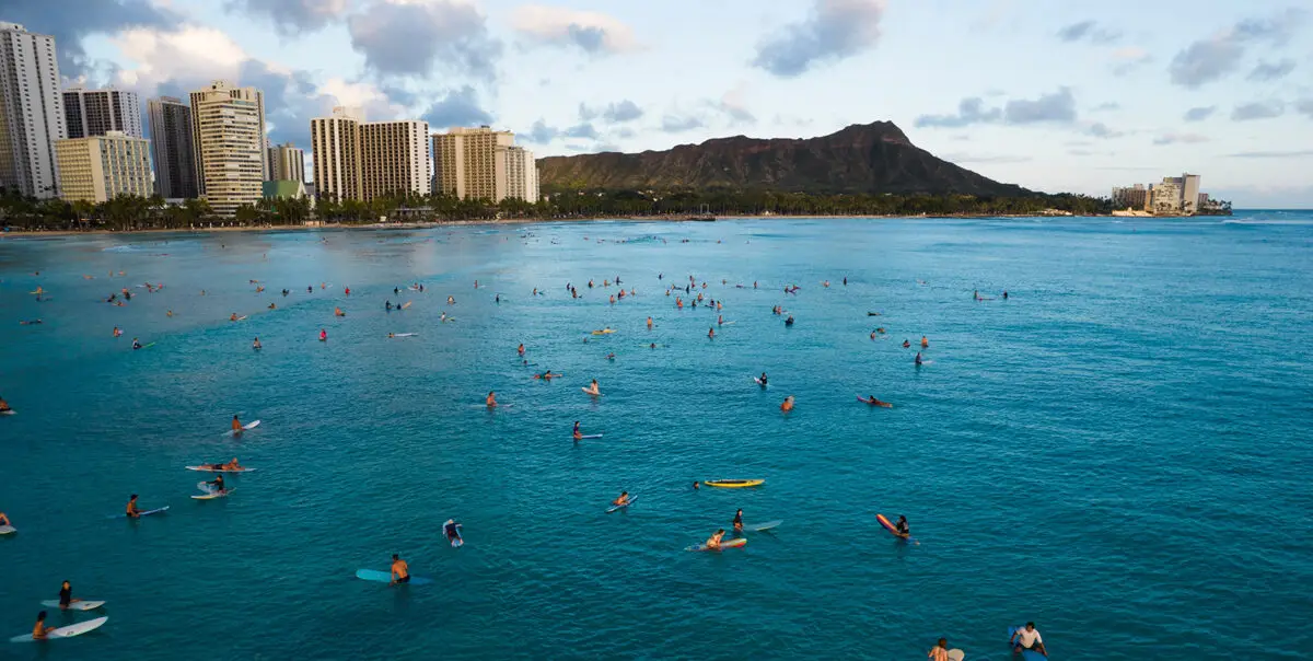 A wide shot of numerous surfers and paddleboarders in the turquoise waters near Waikiki Beach, Honolulu, with the Waikiki skyline and high-rise buildings in the background. Wonder why Waikiki is so iconic? Diamond Head crater is visible in the distance under a partly cloudy sky.