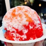 A close-up of a colorful shaved ice dessert in a white bowl, held up against a blurred background. The shaved ice, often touted as the best shave ice in Kauai, is covered in a drizzle of various syrups, creating a vibrant mix of red, orange, and white. A wooden spoon is sticking out from the top.