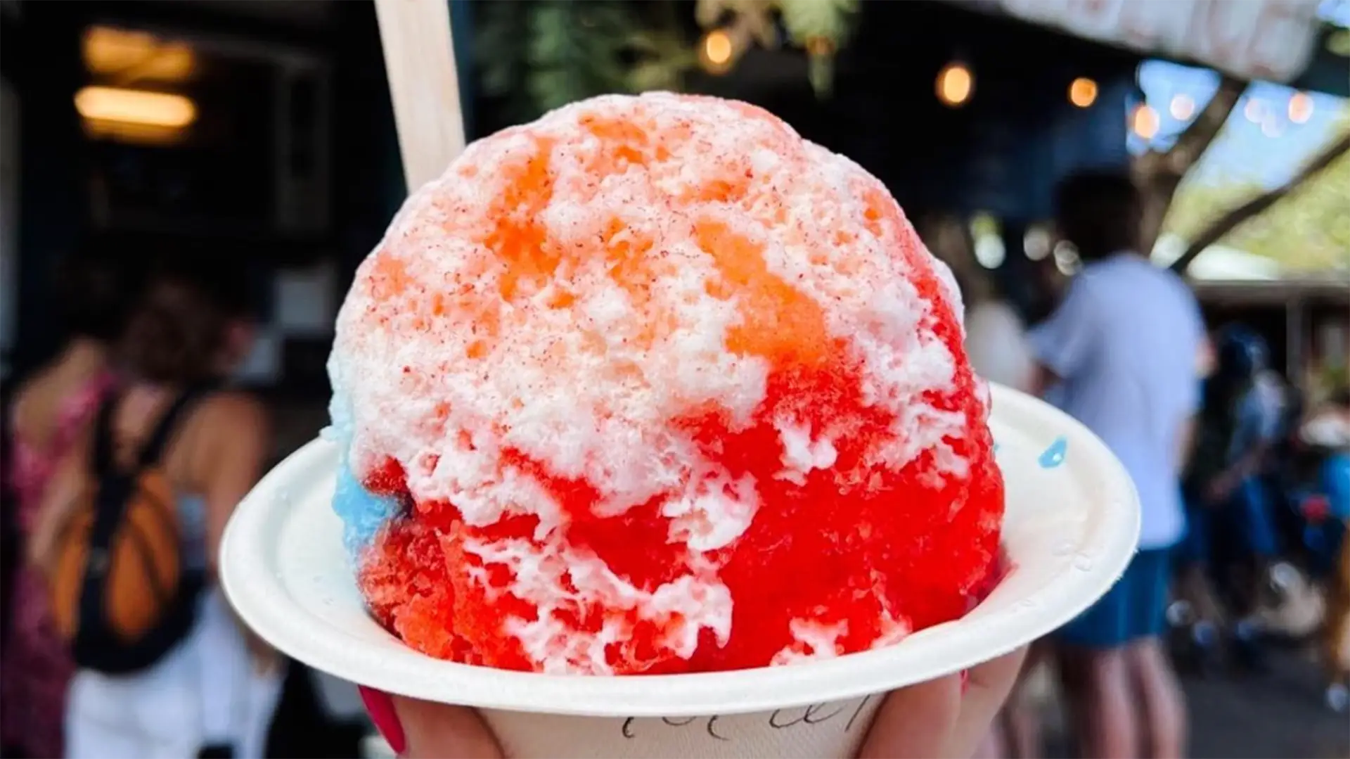 A close-up of a colorful shaved ice dessert in a white bowl, held up against a blurred background. The shaved ice, often touted as the best shave ice in Kauai, is covered in a drizzle of various syrups, creating a vibrant mix of red, orange, and white. A wooden spoon is sticking out from the top.