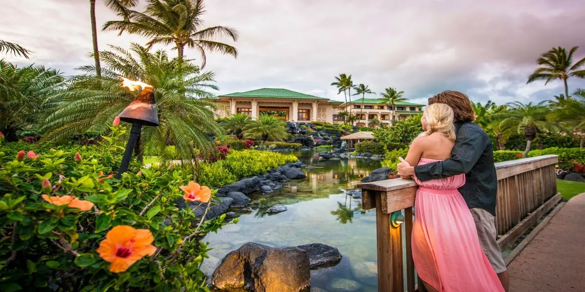 A couple embraces on a wooden bridge adorned with torches, surrounded by lush greenery and vibrant flowers. They overlook a serene pond with a luxurious, tropical resort in the background, offering an idyllic "Where To Stay" experience. The scene is set against a backdrop of palm trees and a partly cloudy sky.