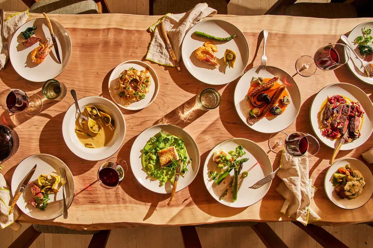 A wooden dining table holds plates of various gourmet dishes, including salads, grilled vegetables, pasta, and meat. Glasses of red wine sit beside the plates. Utensils and napkins are arranged neatly around the setting, reminiscent of one of the best dinner spots in Sonoma County.