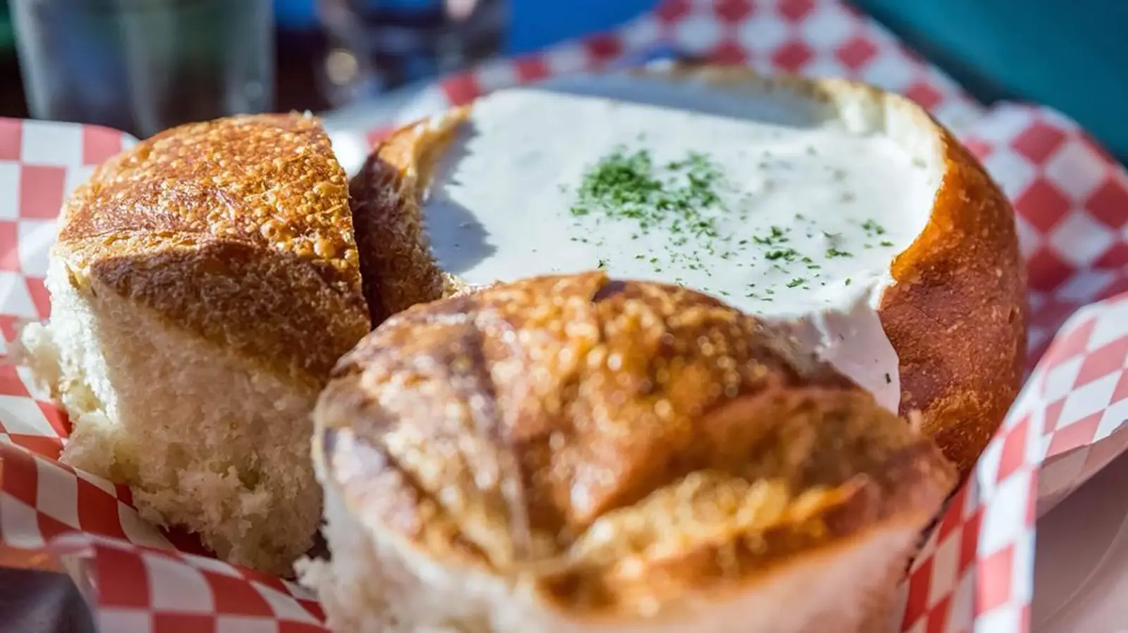 A bread bowl filled with creamy soup is surrounded by chunks of crusty bread. The bowl is placed on a paper liner with a red and white checkered pattern. A sprinkle of herbs garnishes the soup, adding a touch of green to the white, smooth surface—truly the best clam chowder on the Monterey Peninsula.