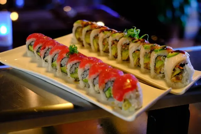 Two platters of sushi rolls are displayed. The roll in the foreground has vibrant red fish slices on top, while the roll in the background features white fish and drizzles of sauce. Both rolls include rice and small green garnishes, served on white rectangular plates—truly the best sushi Kauai has to offer.