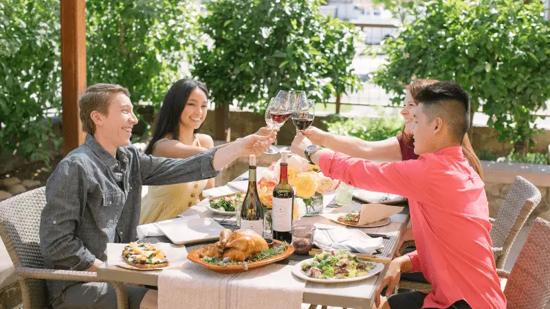 A group of four people sitting at an outdoor dining table, toasting with red wine glasses. The table is set with various dishes, including a roasted chicken, salad, and bottles of wine. They are smiling and appear to be enjoying a pleasant meal together—one of the best things to do in Napa Valley this January.