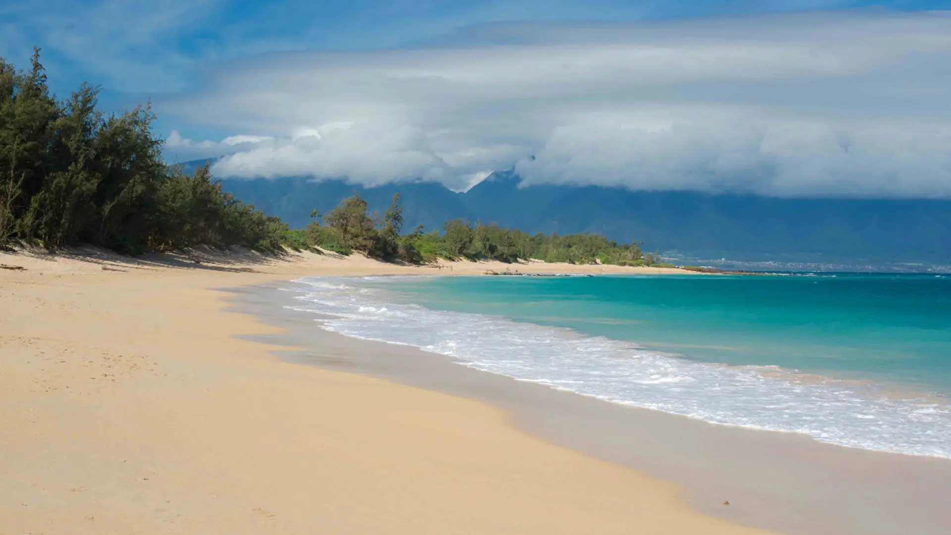 A serene tropical beach with golden sand and turquoise waters. Gentle waves lap at the shore, and lush green trees line the beach. In the background, mountains partially covered by clouds create a stunning contrast against the clear blue sky, making it one of Maui's best family beaches.